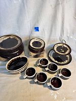 Wide view of Pfaltzgraff stoneware pieces including stacked plates, mugs, four oval bowls, and two-tier serving stand with brown drip glaze