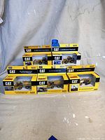 Five boxed CAT 1:87 scale diecast construction vehicle models arranged in two rows, showing boxes and branding