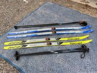 Full view of two pairs of kids cross country skis, blue Atomic and yellow Fischer, along with two pairs of black ski poles on a fabric surface outdoors.