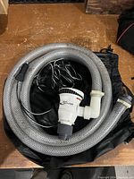 Full bilge pump with coiled braided clear tubing and attached cable laid in black drawstring bag on wooden surface.
