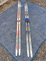 Two pairs of adult cross-country skis laid on a blue textured mat: the Fischer RCS pair 190 cm on left and Atomic ARC pair 185 cm on right, both with SNS Profil bindings.
