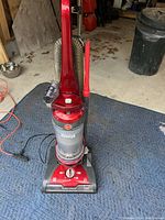 Front view showing upright vacuum cleaner with attached hose and crevice tool on blue padded mat with background of garage floor and some equipment.