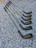 Photo showing full set of seven left handed Cleveland golf irons laid on ground, grip ends towards top and club heads towards bottom. Clubs numbered 3,4,5,6,8 and a pitching wedge.