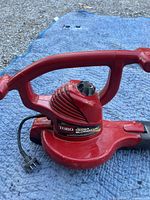 Side view of Toro Ultra Blower Vac showing red housing, handle and attached electric cord with plug on blue surface.