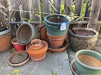 Assorted planter pots shown stacked and grouped together outdoors on concrete, including terracotta and glazed pots.