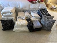 Wide view of all bedding items arranged on floor in front of couch, showing white quilts, folded sheets, variety of plush blankets in colors including black, gray, plaid, plus zebra and wavy patterned pillows.