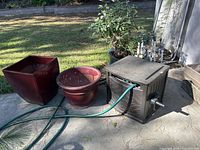 Photo of two burgundy glazed terracotta planters (one round and one square) alongside a brown Suncast Hose House with an attached green hose, placed on outdoor pavement.