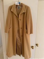 Front view of the camel hair coat hanging on a door, showing full length, collar, button closure, and pockets.