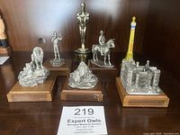 Five pewter figurines on wooden bases displayed along with two plastic souvenirs: a golden award figure and a yellow Eiffel Tower.
