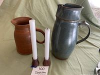 Blue/grey glazed stoneware pitcher, rust colored stoneware pitcher, two brown stoneware candle holders with white candles, all on a light green fabric background.