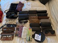 Full view of assorted sunglasses, readers, and multiple eyewear cases including branded hard cases and fabric pouches laid out on a beige surface.
