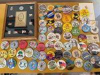 Full lot of assorted colorful vintage pins and buttons spread on a table, including many round event and novelty pins and a framed pin set in the top left corner.