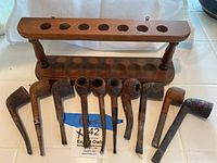 Photo showing the full lot with 10 wooden pipes arranged in front of a wooden pipe holder with 10 slots for pipes, featuring turned wooden supports.