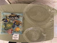 Three fish-themed glass serving dishes shown on green tablecloth: large clear glass fish platter, smaller clear glass fish platter, and painted square glass dish with colorful fish