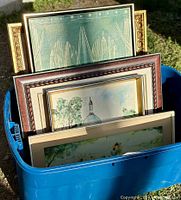Blue bin holding multiple framed pieces of art, including a framed watercolor and painting visible at the front.