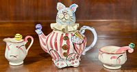 Front view of the whimsical bunny rabbit shaped teapot, sugar bowl with spoon, and milk jug arranged on a wooden surface.