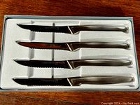 Set of four J.A. Henckels steak knives in original box with fitted white foam insert. The knives have polished stainless steel handles and serrated blades.