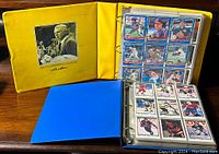 Two large card albums, yellow and blue, displayed open with baseball and hockey cards in clear sleeves. Yellow album shows a page of nine cards including various baseball players.