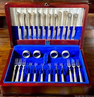 Full view of the wooden Birks chest open showing 12 ornate knives in the upper compartment and 4 napkin rings with 12 forks in the lower compartment on blue velvet lining.