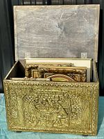 Brass repousse firewood box with hinged lid open showing embossed outdoor scene with people around table.
