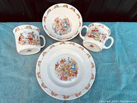 Photo of all four dish pieces: two mugs, one bowl, one plate laid on blue cloth background showing the Bunnykins illustrations and bunny borders