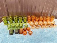 Wide view of all pressed glass green and amber tumblers, stemware, and salts arranged on blue cloth.
