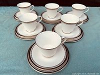 Six white bone china cups each paired with a matching saucer, all decorated with the Royal Doulton Sarabande pattern of silver detailed banding on rim and base, displayed on blue cloth.