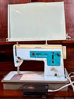 The full sewing machine shown on a wooden surface with the original beige lid case behind it and the black foot pedal in front lying flat.