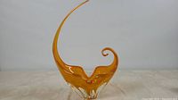 Front angled view of amber art glass sculpture showing the curved tall neck and curled tip, glossy finish and clear glass base accents