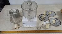 Full view of Cuisinart food processor components including base unit, large and small bowls, lids, blades, discs, and accessories arranged on a table.
