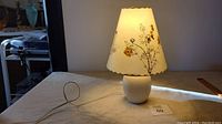 Full view of the lamp on a table, showing white ceramic base and illuminated floral shade.