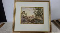 Framed watercolour artwork showing the entire painting with a double matted border and wooden frame with gold trim. Image captures the general composition of village in distance with figures in foreground.