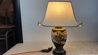 Full view of the lamp including ceramic body, wooden base, beige pergola lampshade, and electric cord with plug