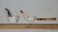 Overview of three smoking-related items: two clay pipes (one skull face pipe with silver band, one simpler clay pipe) and one amber colored cigarette holder.