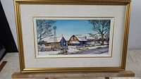 Framed oil painting depicting a winter rural scene with barns and a windmill, labeled and signed by artist Sam Saunders.