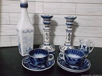 Full group view of Delft blue and white collection including candlesticks, bottle, creamer, and cups with saucers arranged on black surface against tiled white background.