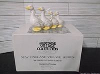 Four white and yellow porcelain calling bird figurines neatly aligned on top of the original box labeled Heritage Village Collection, New England Village Series, McGrebe Cutters & Sleighs, handpainted porcelain figurines.