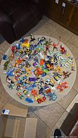 Wide view of assorted colorful vintage action figures scattered on a round beige carpet including superheroes, movie characters, and cartoon figures.