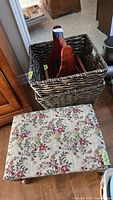 Photo showing large and small dark brown wicker baskets stacked, wooden magazine rack inside larger basket, floral brocade foot stool in front on wood floor