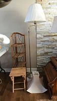 Full view of floor lamp with white shade, wooden folding stool, and wooden corner shelf unit