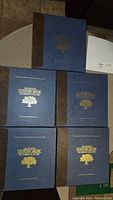 Five hardcover boxed volumes arranged on a surface, showing front covers with blue fabric, brown spine, gold lettering, and embossed tree logo.