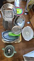 Overview photo showing stacks of metal bakeware pans, cooling racks, silicone cutting boards, plastic bowls and trays, strainers, and other kitchen items arranged on the floor.