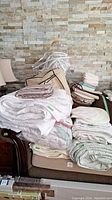 Stack of assorted queen size bedding linens including sheets, mattress protectors, folded linens piled on chair