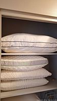 Four white bed pillows stacked on a shelf. Three pillows are memory foam with quilted covers, and one pillow is polyester filled with white fabric featuring a black trim.