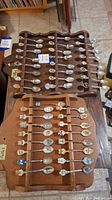 Two wooden souvenir spoon racks, each holding multiple souvenir spoons with colorful crests and designs, displayed on a wooden surface.