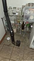 Metal wire shelving unit with glass jars, bottles, and a plastic drawer organizer in a basement corner with black plumbing pipes nearby