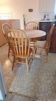 Photo showing maple round pedestal table with four wooden spindle-back chairs around it on patterned floor.