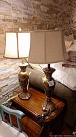 Two metal table lamps with beige fabric shades resting on a wooden surface against a stone wall background.
