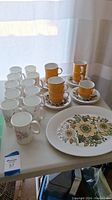 Photo of all cups, saucers, mugs, and serving plate arranged on a table, showing the variety and quantity of the lot.