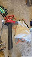Full side view of red Toro electric leaf blower/vacuum with attached white cloth collection bag and orange extension cord laid on a rough concrete floor.